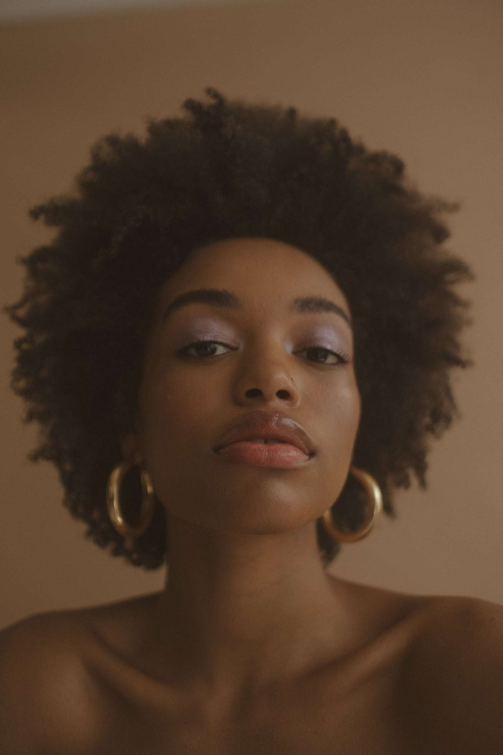 A woman with a full afro and gold hoops, against a warm tan wall, from Deun Ivory’s campaign for Glossier