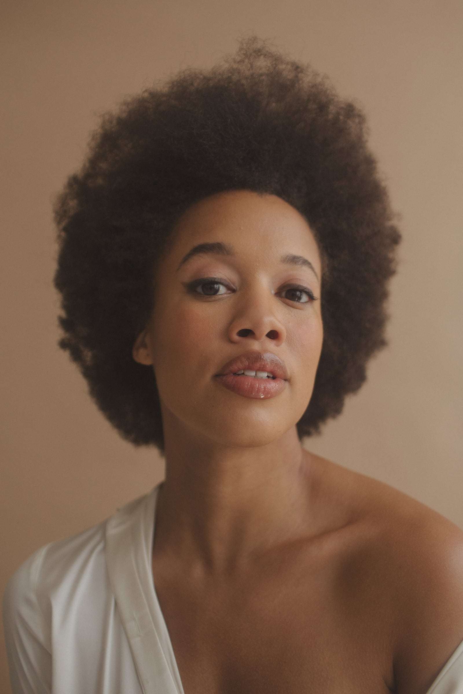 A woman with a full afro in a white top, against a warm tan wall, from Deun Ivory’s campaign for Glossier