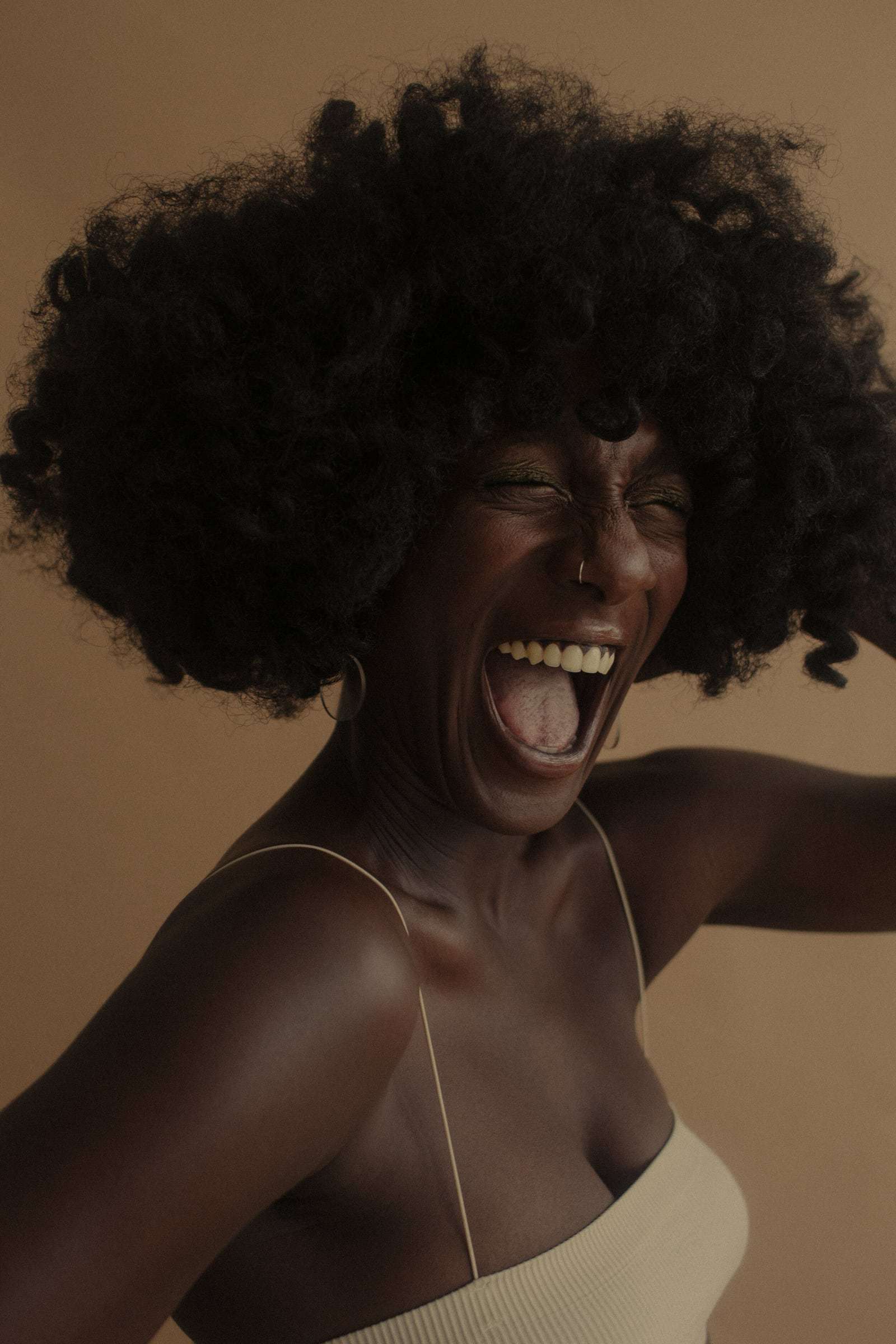 A woman laughing, head thrown back, against a warm tan wall, from Deun Ivory’s campaign for Glossier