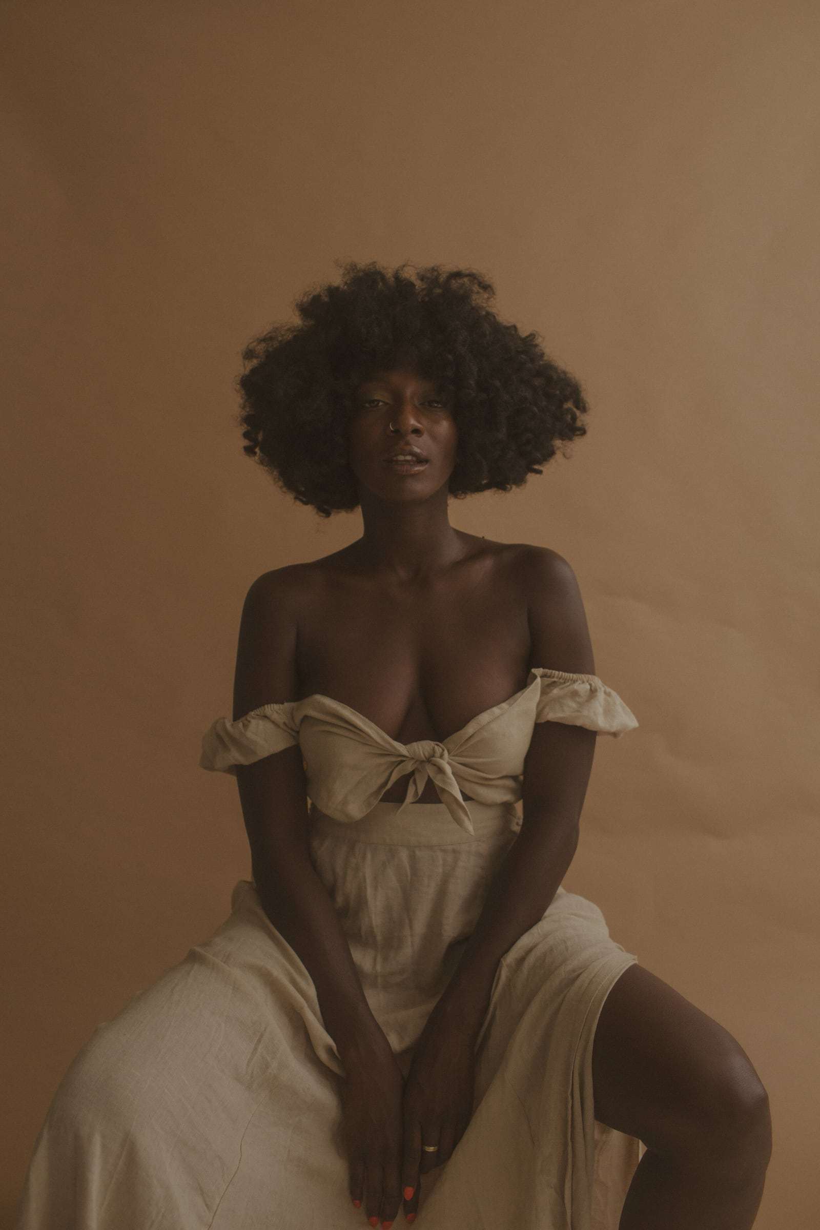 A woman in an off-shoulder cream dress, seated against a tan backdrop, from Deun Ivory’s campaign for Glossier