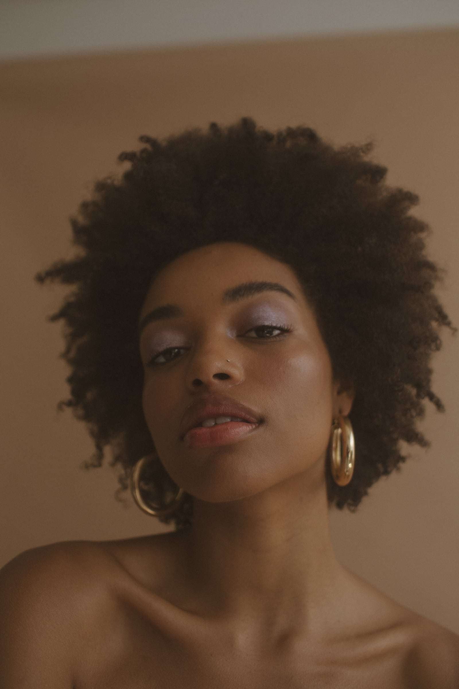 A woman with a full afro and gold hoops looking down at the camera, from Deun Ivory’s campaign for Glossier