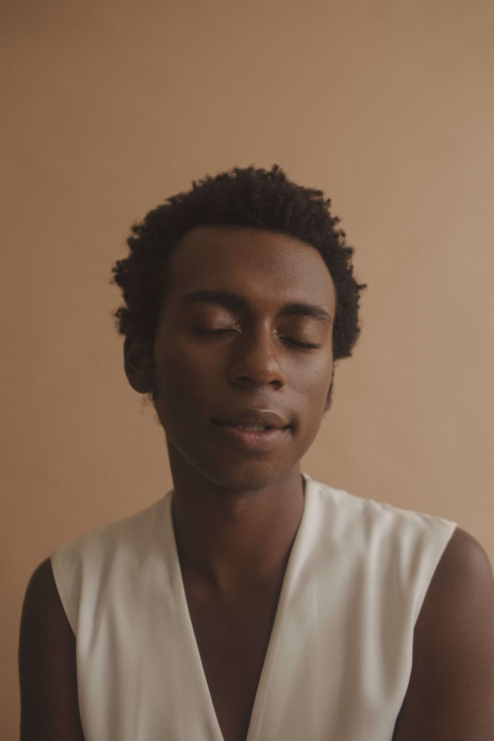 A man with his eyes closed, in a white vest, against a tan wall, from Deun Ivory’s campaign for Glossier