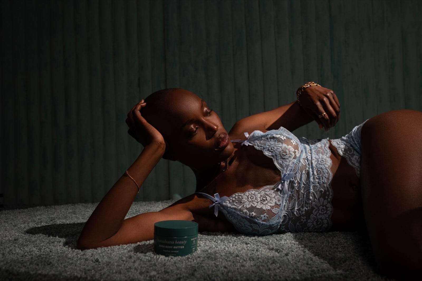 A woman lying on a dark green carpet beside a jar of body butter, from Deun Ivory’s campaign for Hanahana Beauty at ULTA