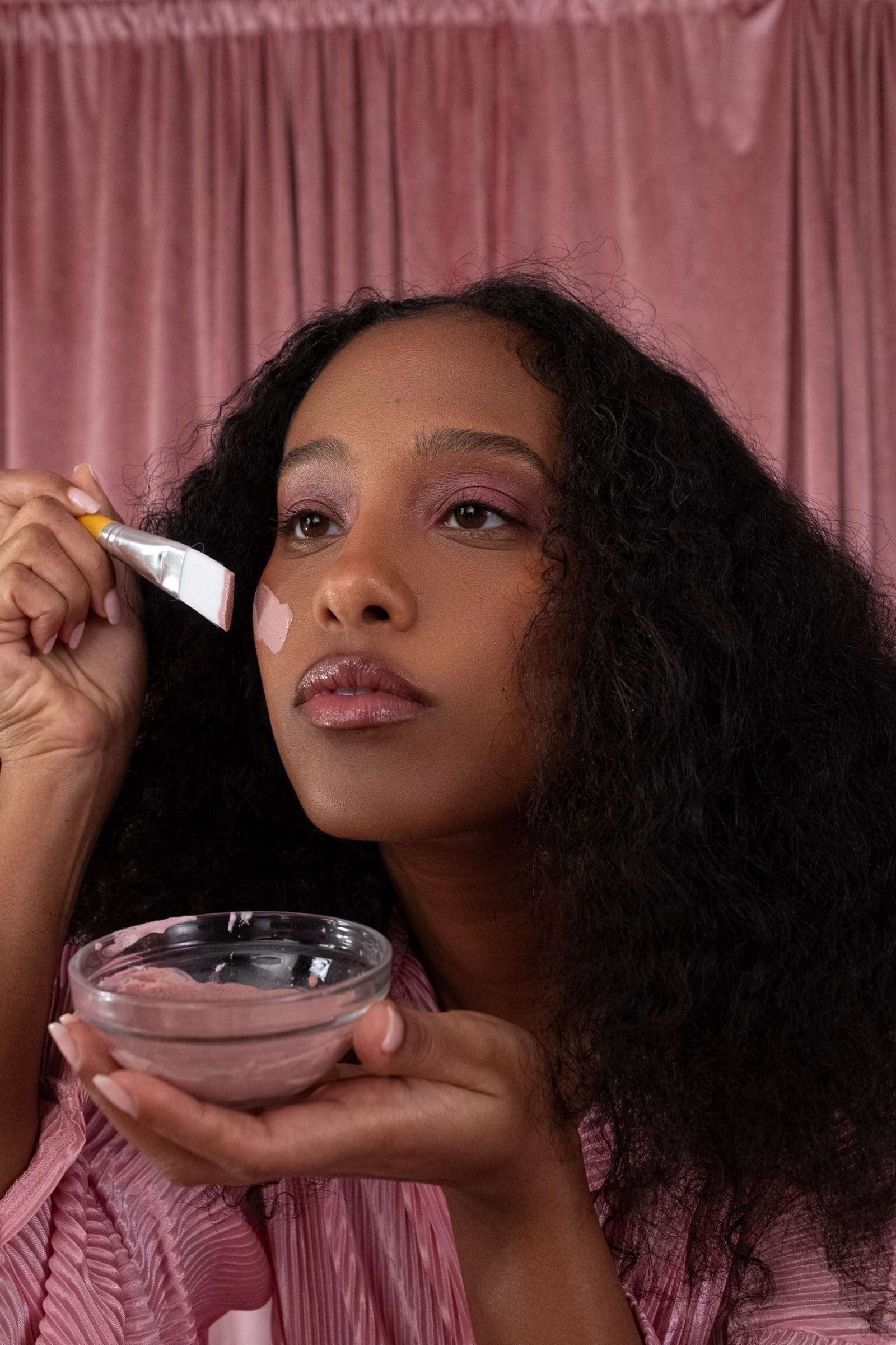 A woman in pink touching cream to her cheek from a glass bowl, before a pink curtain, from Deun Ivory’s campaign for Hanahana Beauty at ULTA