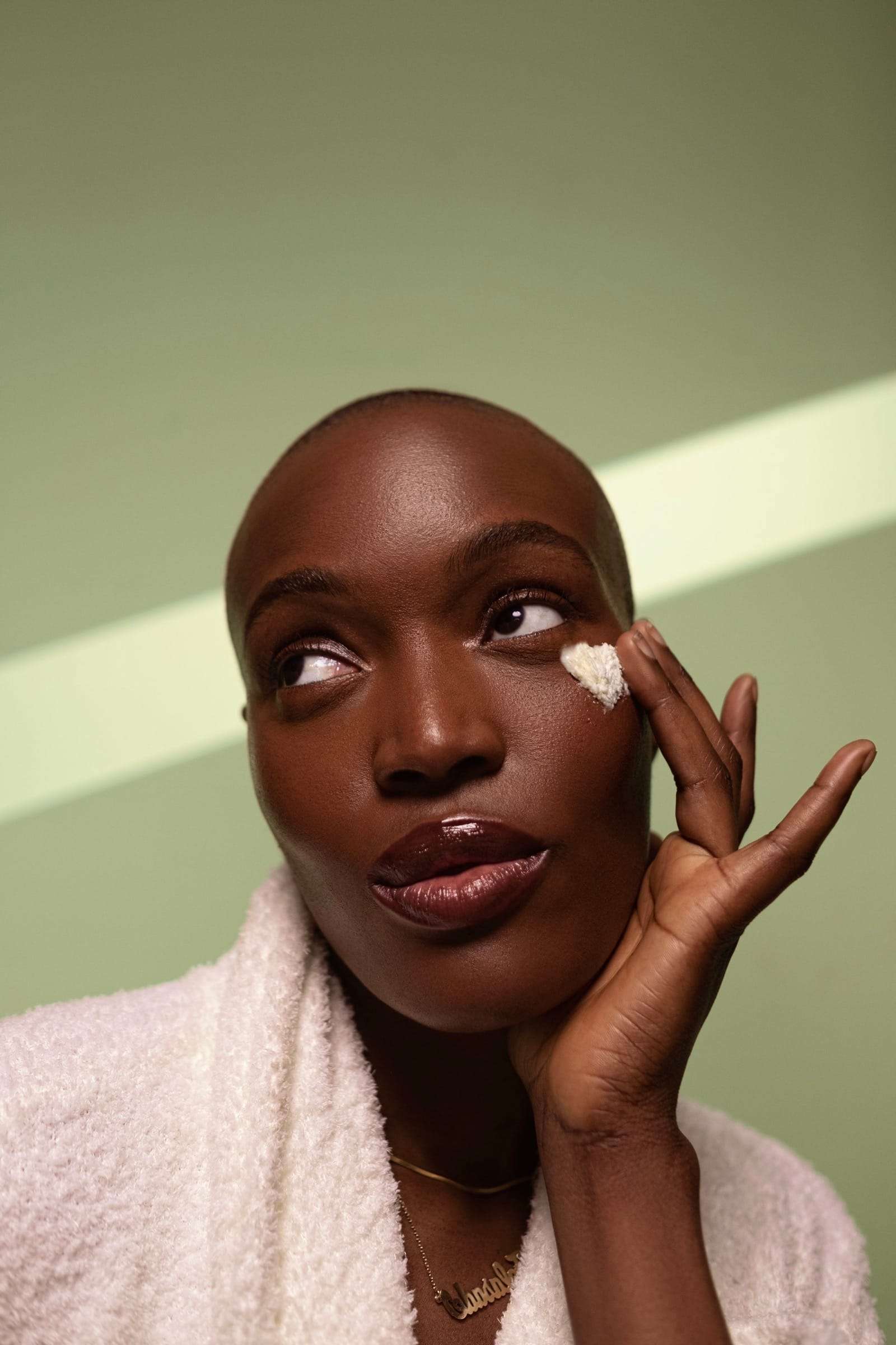 A woman in a white robe touching cream to her cheek, against a sunlit green wall, from Deun Ivory’s campaign for Hanahana Beauty at ULTA