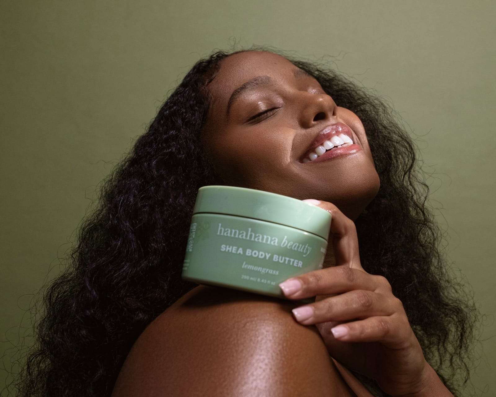 A smiling woman holding a jar of shea body butter to her shoulder, from Deun Ivory’s campaign for Hanahana Beauty at ULTA
