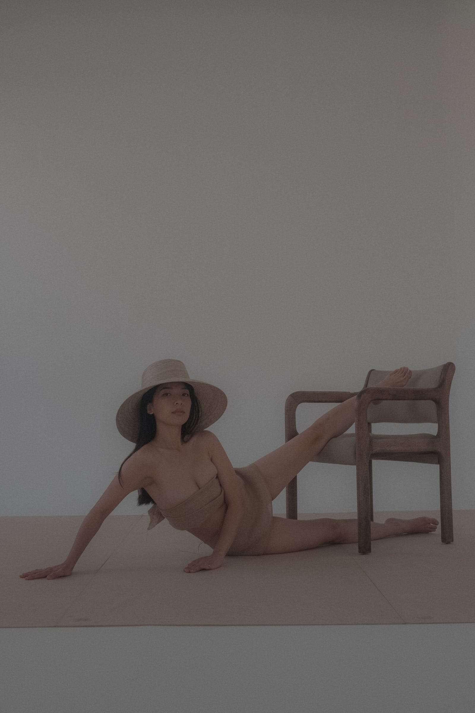 A woman in a straw hat stretched on the floor of a white studio beside a wooden chair, from Deun Ivory’s campaign for Honeynsilk