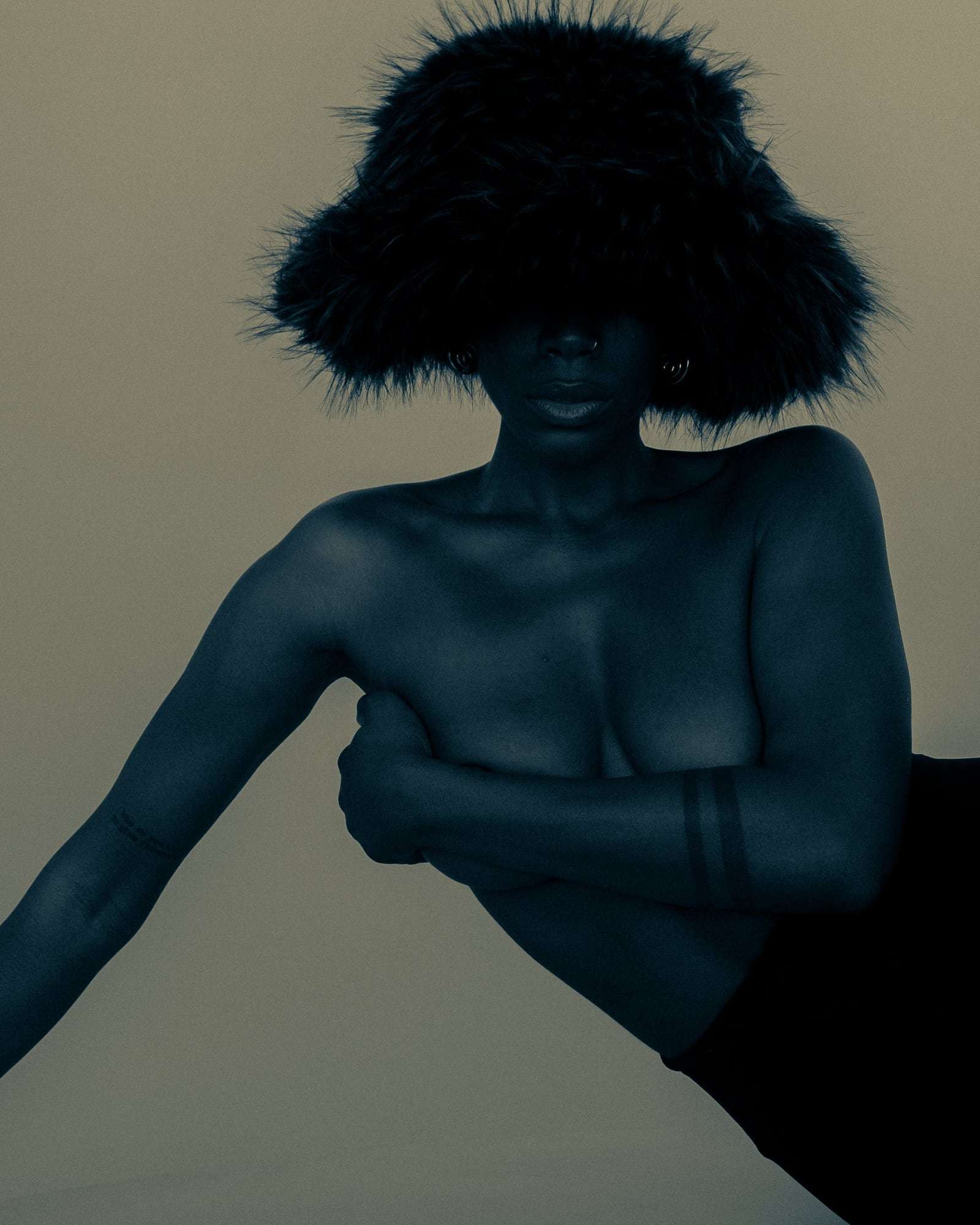 A figure in a furry hat, arms folded across the chest, in blue light, from Deun Ivory’s campaign for Youth To The People